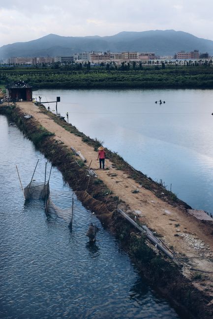 A fisherman walks along a narrow path by a lake, framed by mountains and a distant town.