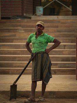 Man wearing green shirt and cap standing with shovel on urban stairs in Bhubaneswar, India.