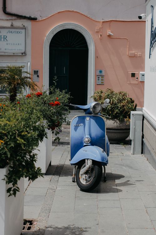 Free A classic blue Vespa scooter parked on a sunny day amid vibrant potted plants. Stock Photo