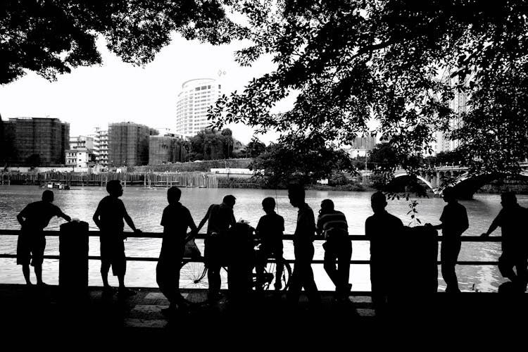 Silhouettes Of People Standing In Front Of A City River Railing