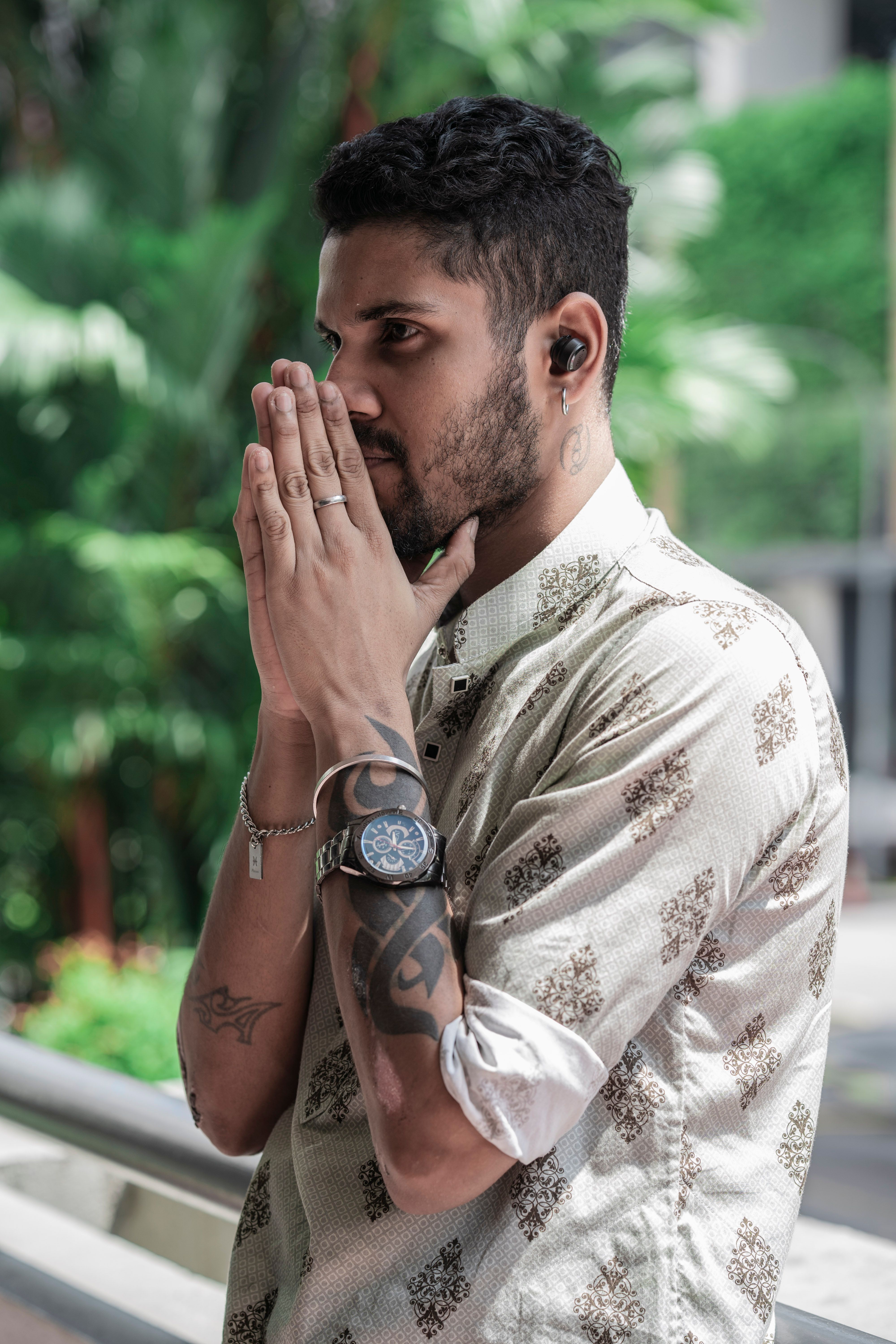 Man with tattoos and watch in thoughtful pose outdoors, tropical background.