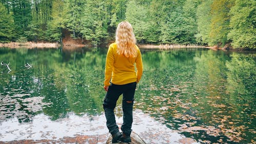 Beautiful young blonde girl sitting on a fallen tree and looking at the spring lake