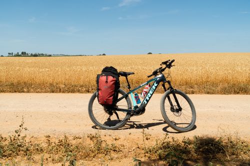 Free A mountain bike with a red backpack parked on a rural dirt road next to expansive golden fields under a clear blue sky. Stock Photo