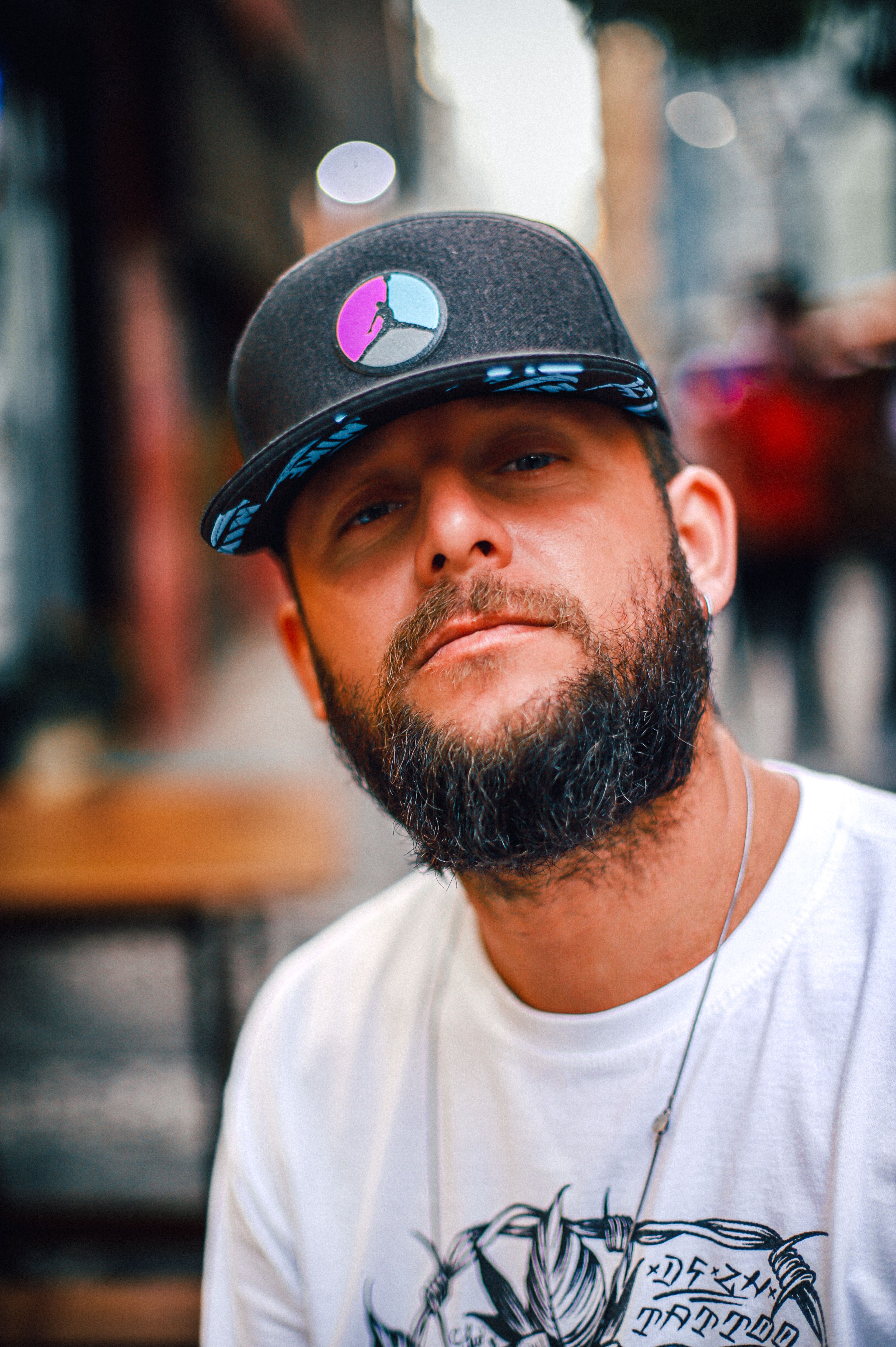 Portrait of a bearded man wearing a stylish cap, taken outdoors in a vibrant urban environment.