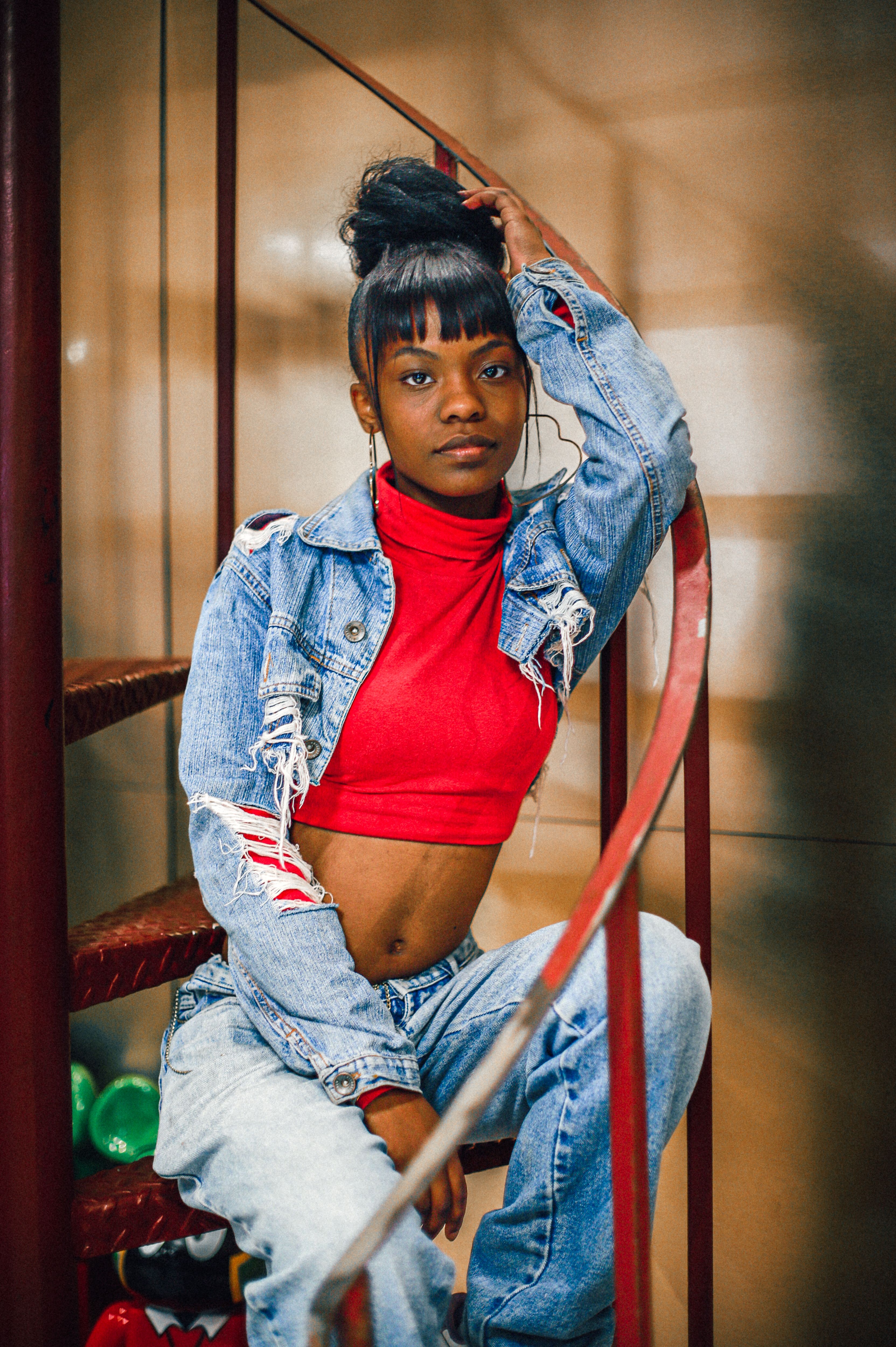 Stylish woman in ripped denim jacket and red top posing confidently on spiral stairs.