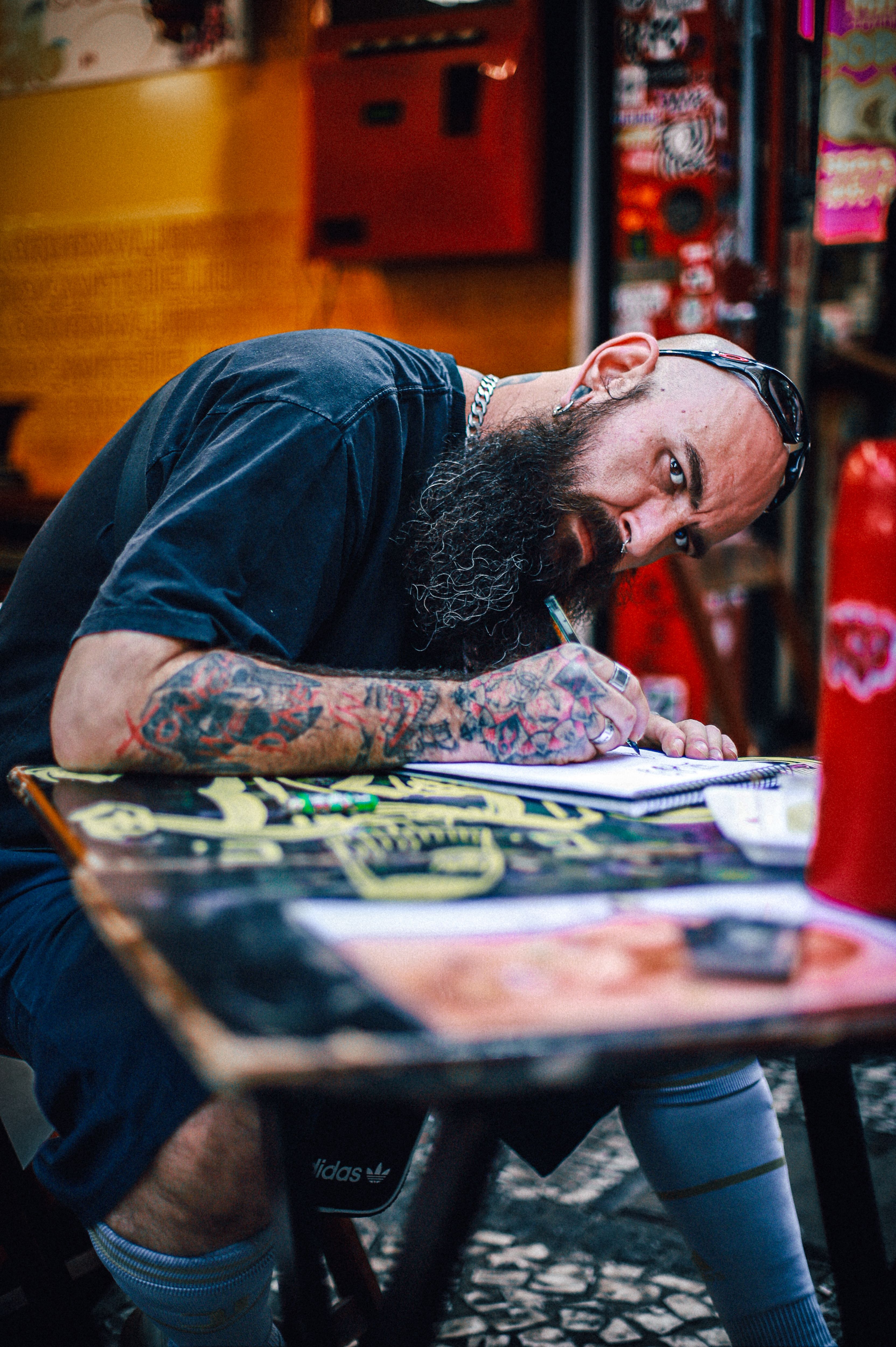 Bearded man with tattoos writing at an urban outdoor cafe table.