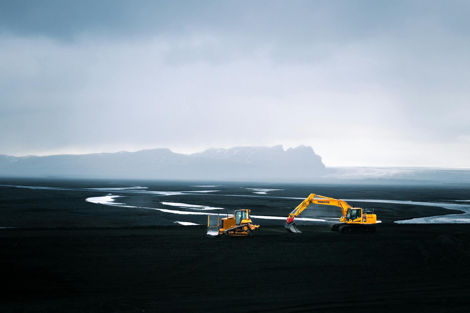 An aerial shot capturing a bulldozer and excavator in a vast mountain valley landscape under a cloudy sky.