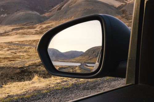 Free View of a winding road through mountainous terrain, captured in a car's side mirror at sunset. Stock Photo