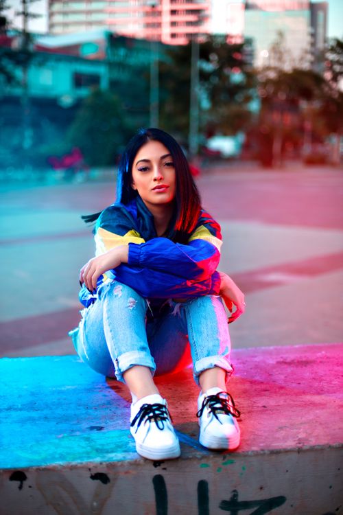 Free Stylish woman in colorful jacket and ripped jeans, sitting outdoors with vibrant cityscape backdrop. Stock Photo