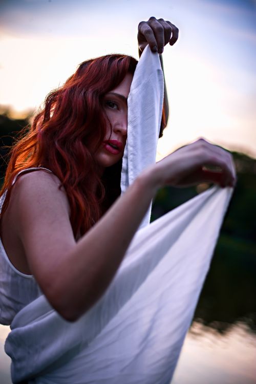 Free A red-haired woman enveloped in white cloth stands by a tranquil lakeside at twilight. Stock Photo