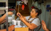 Mujer atiende a clientes en un puesto de comida concurrido mientras sostiene un teléfono.