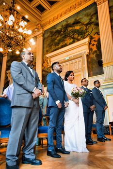 A couple stands at an indoor wedding ceremony, elegantly dressed with guests and chandeliers.