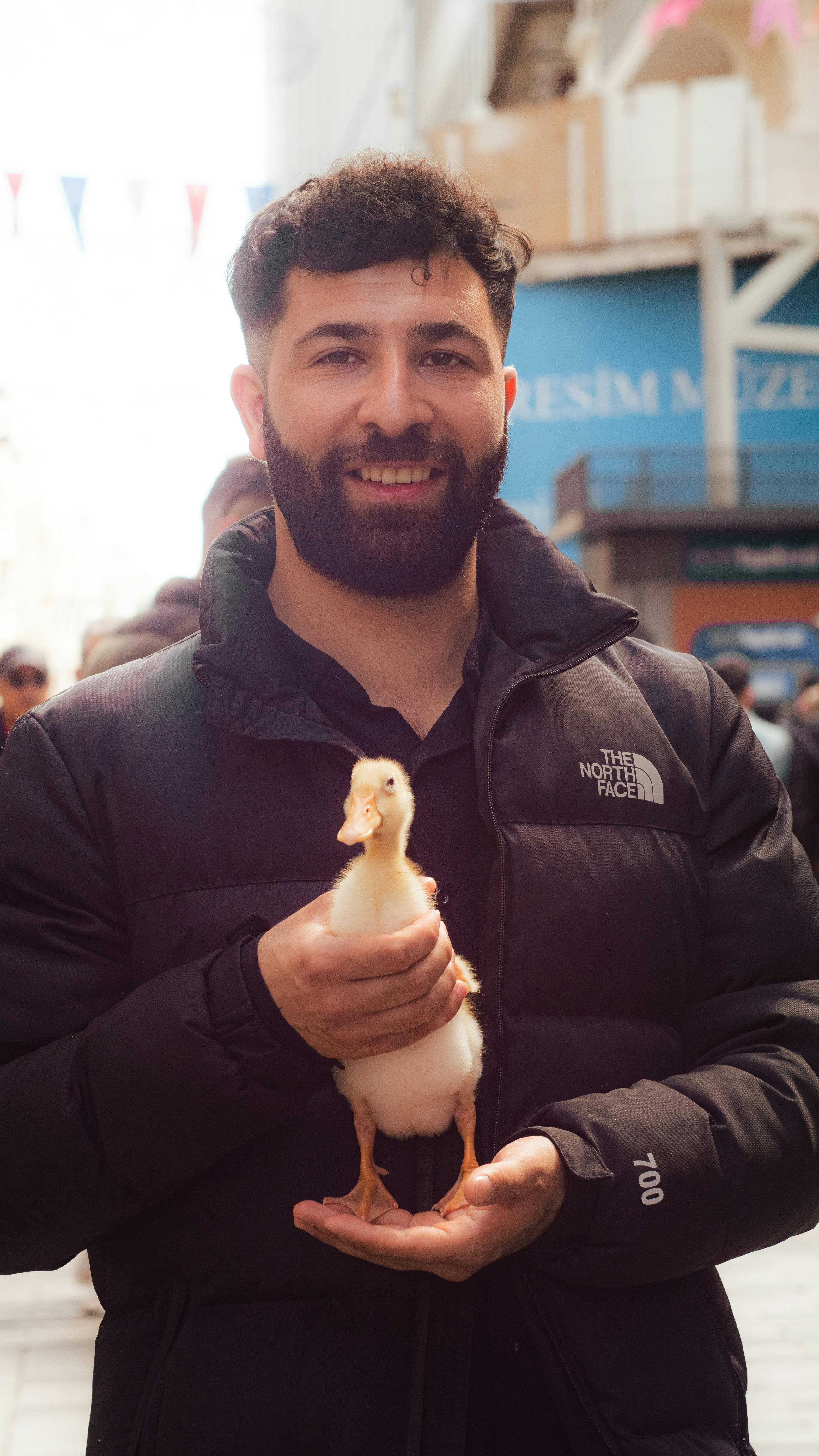 Smiling man in a black jacket holding a small duck outdoors on a sunny day.