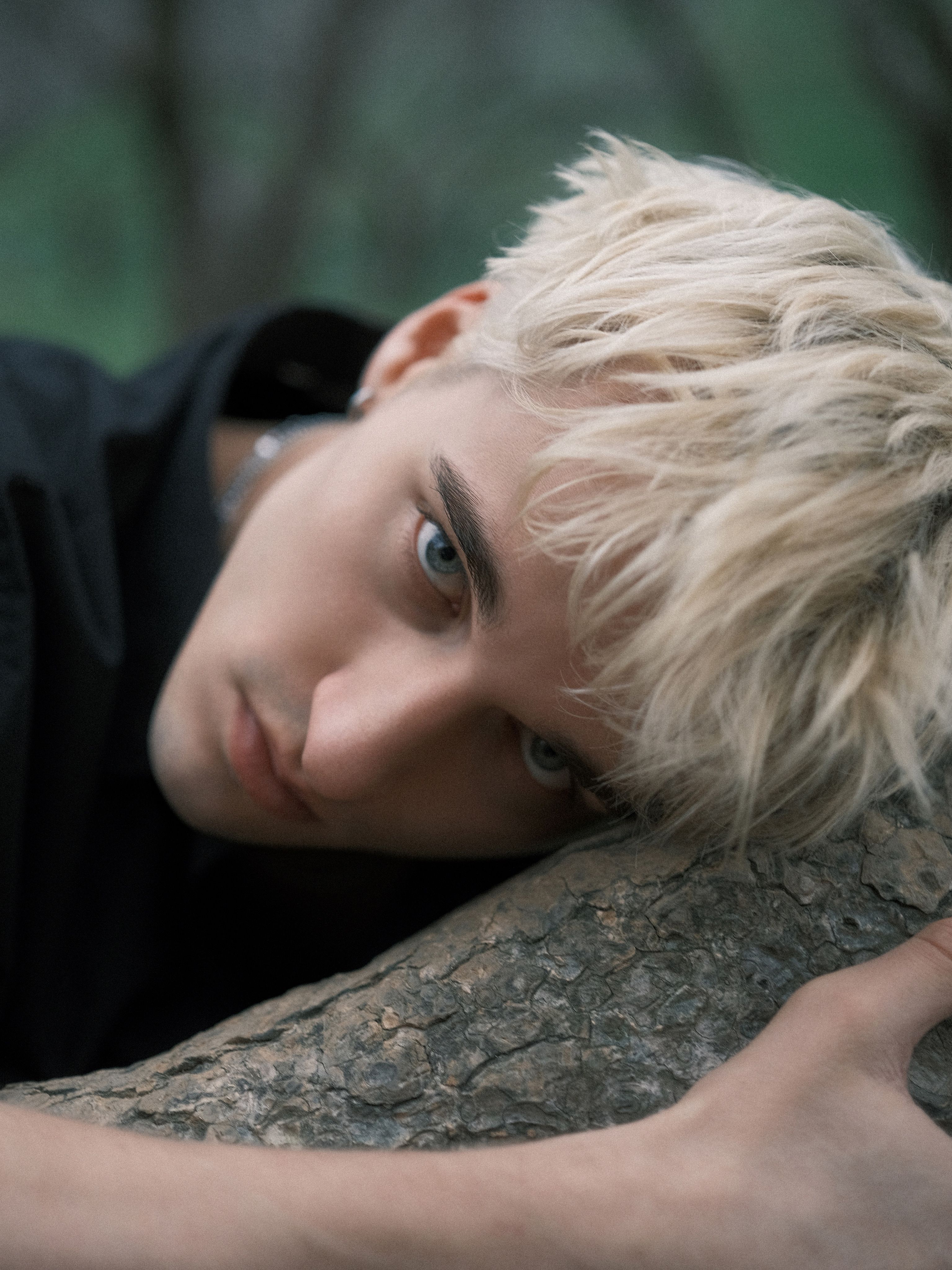 Close-up portrait of a young man with blonde hair leaning against a tree trunk.