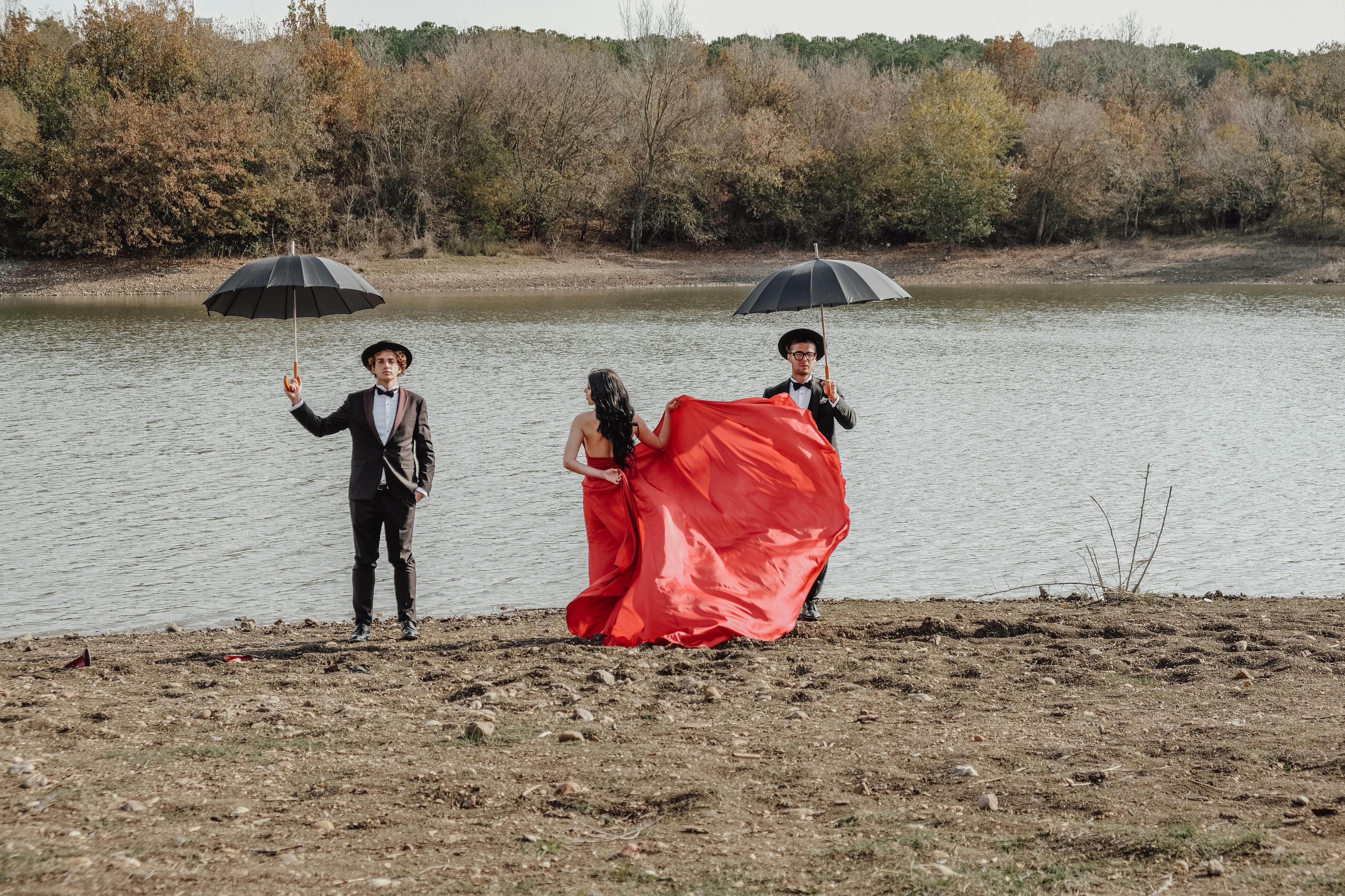 Stylish trio in formal attire with umbrellas near a tranquil lake setting.