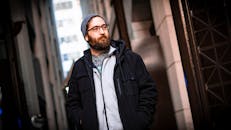 一名留着胡须、头戴无檐帽、身穿夹克的男子自信地站在城市小巷里