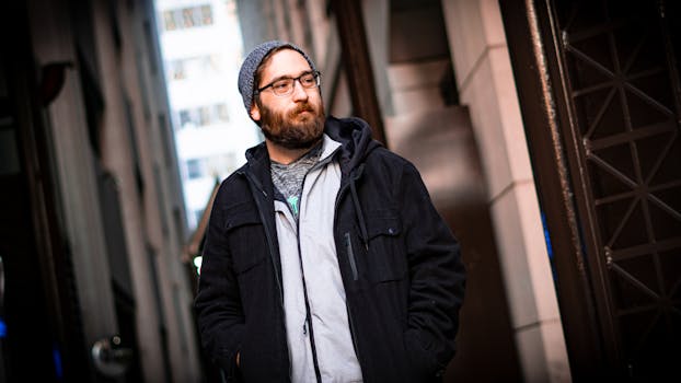 一名留着胡须、头戴无檐帽、身穿夹克的男子自信地站在城市小巷里