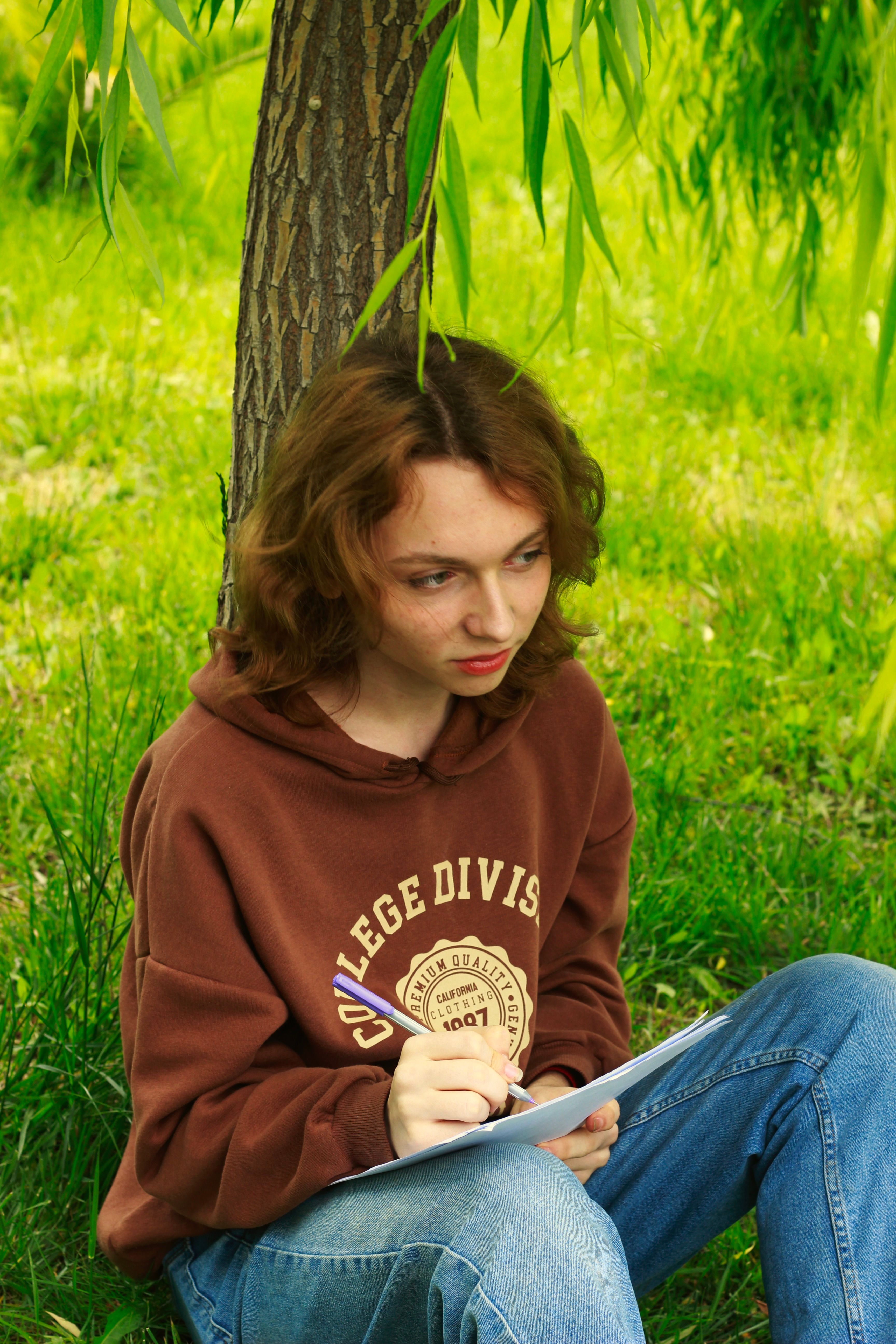 A young redhead woman sits under a lush tree, writing in a notebook outdoors.