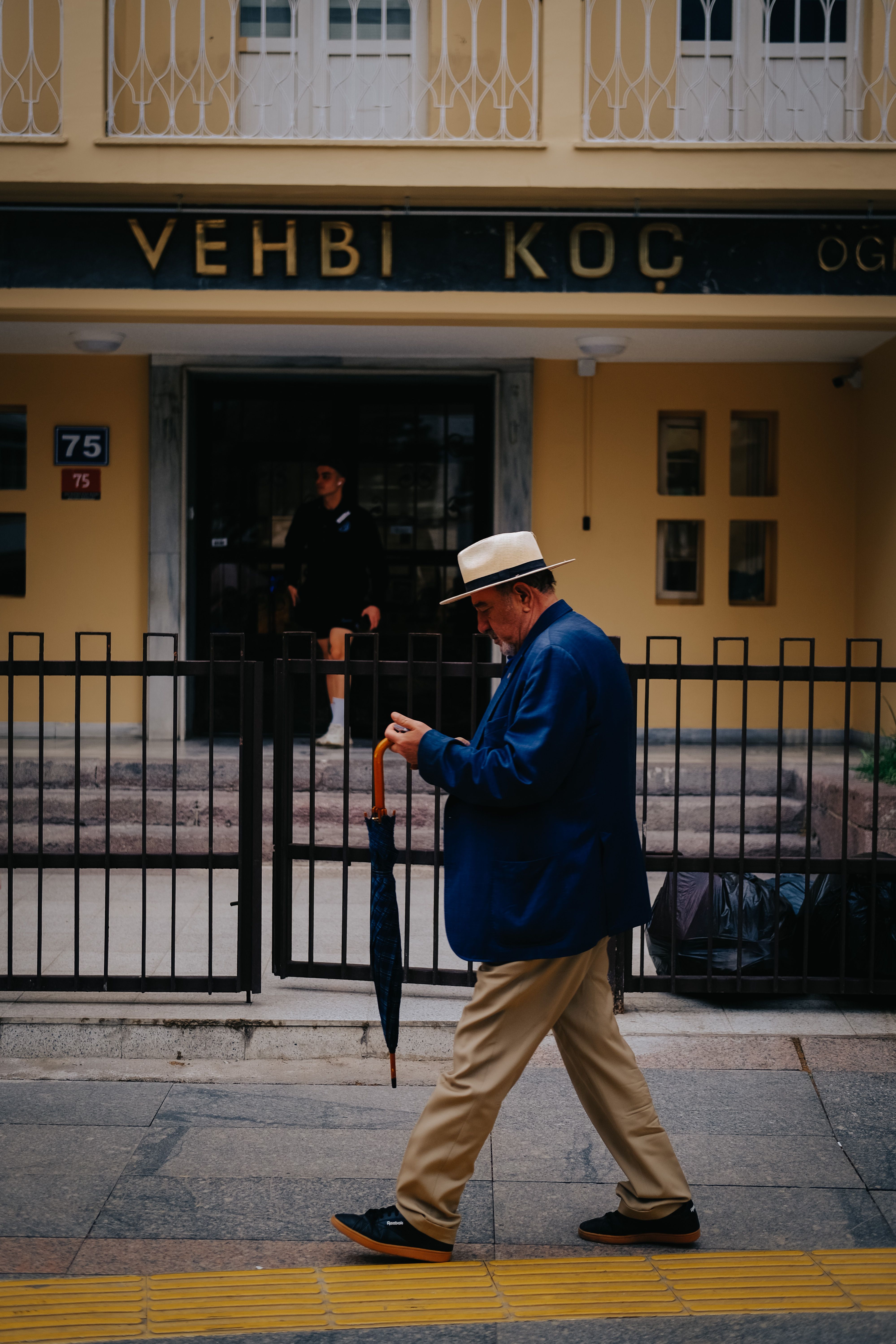 Senior man with a white hat and blue blazer walking down Ankara sidewalk holding an umbrella.
