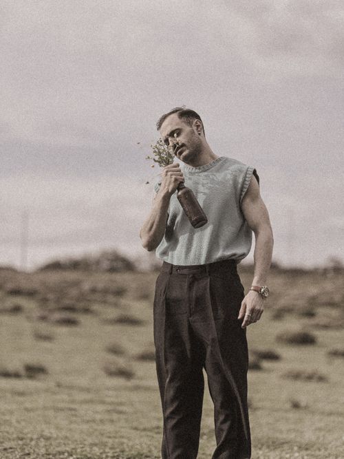 Free Man standing outdoors holding a bottle with flowers, wearing a tank top in a grassy field. Stock Photo
