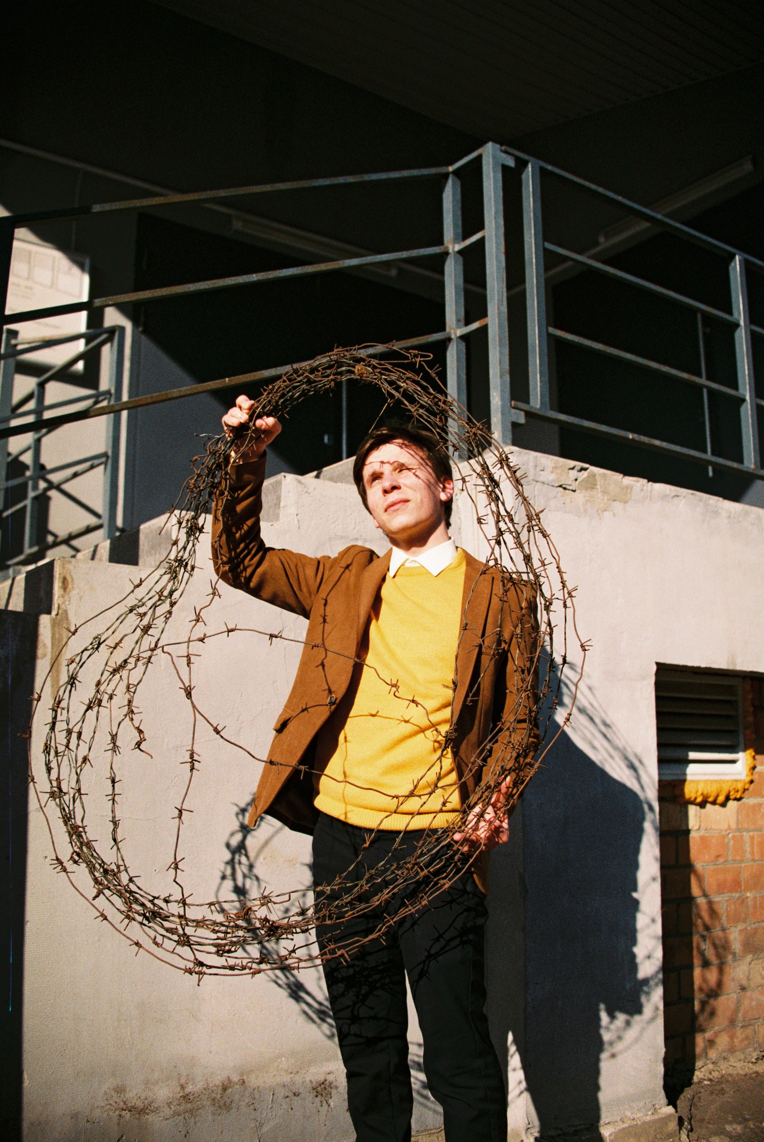 A stylish man poses with barbed wire in sunlight, against an industrial wall.
