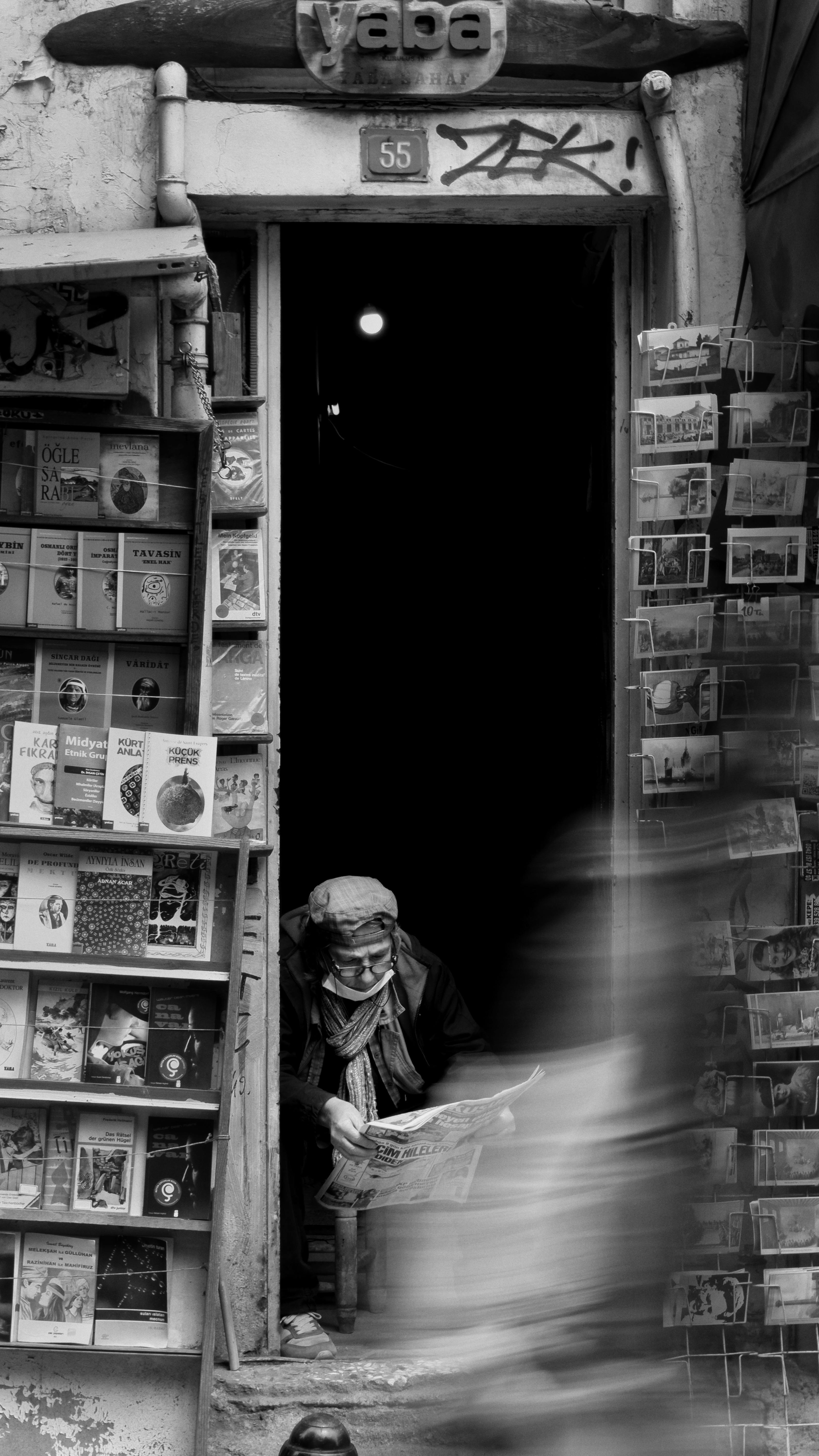 Black and white photo of an elderly man reading in a doorway surrounded by books.