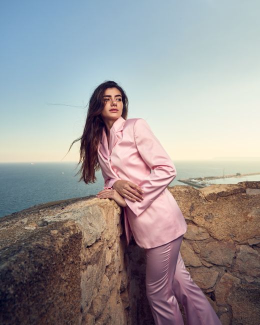 Fashion portrait of a woman in a pink suit leaning against a coastal stone wall with sea view.