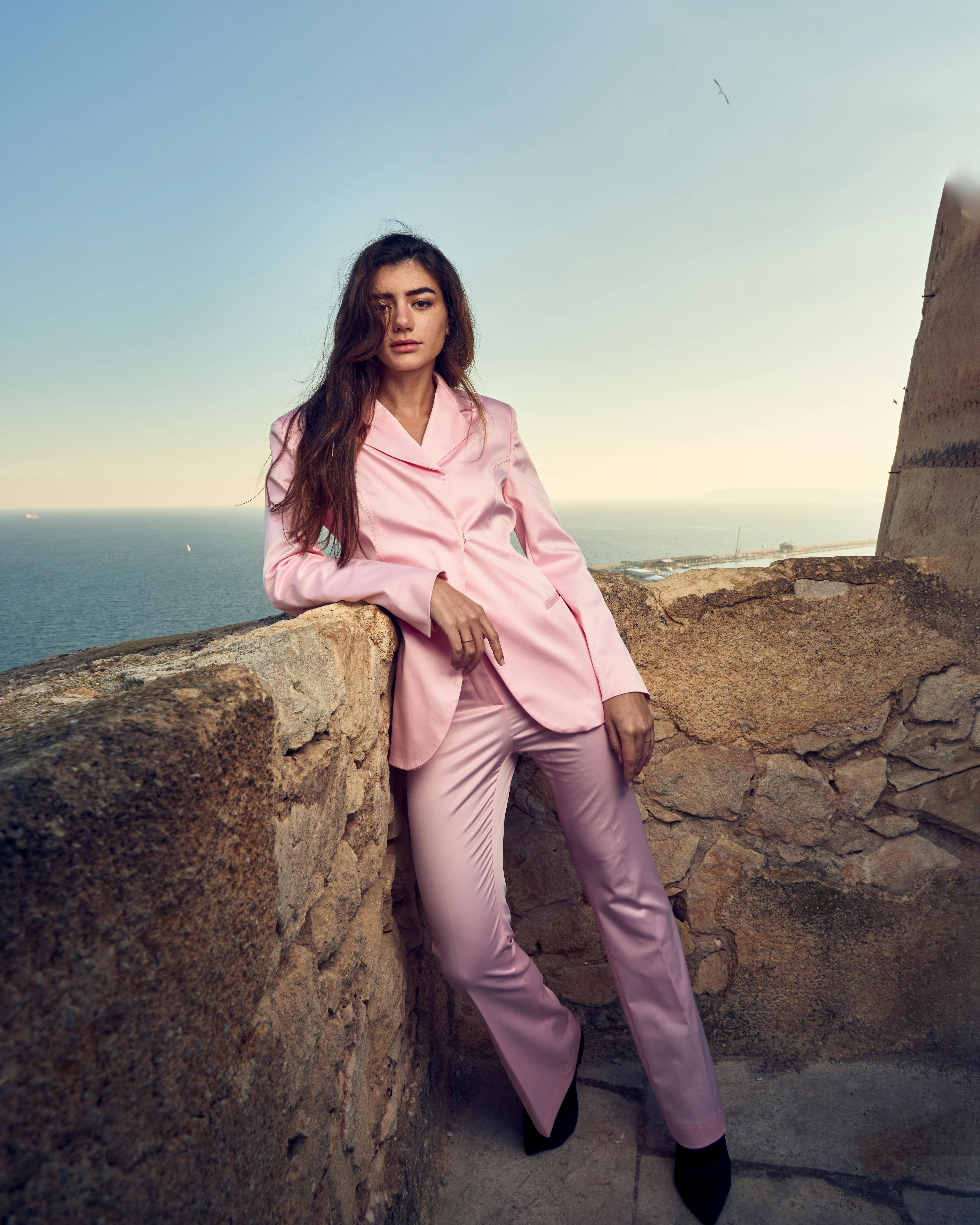 Elegant woman poses in a pink suit against a coastal stone wall at sunset.