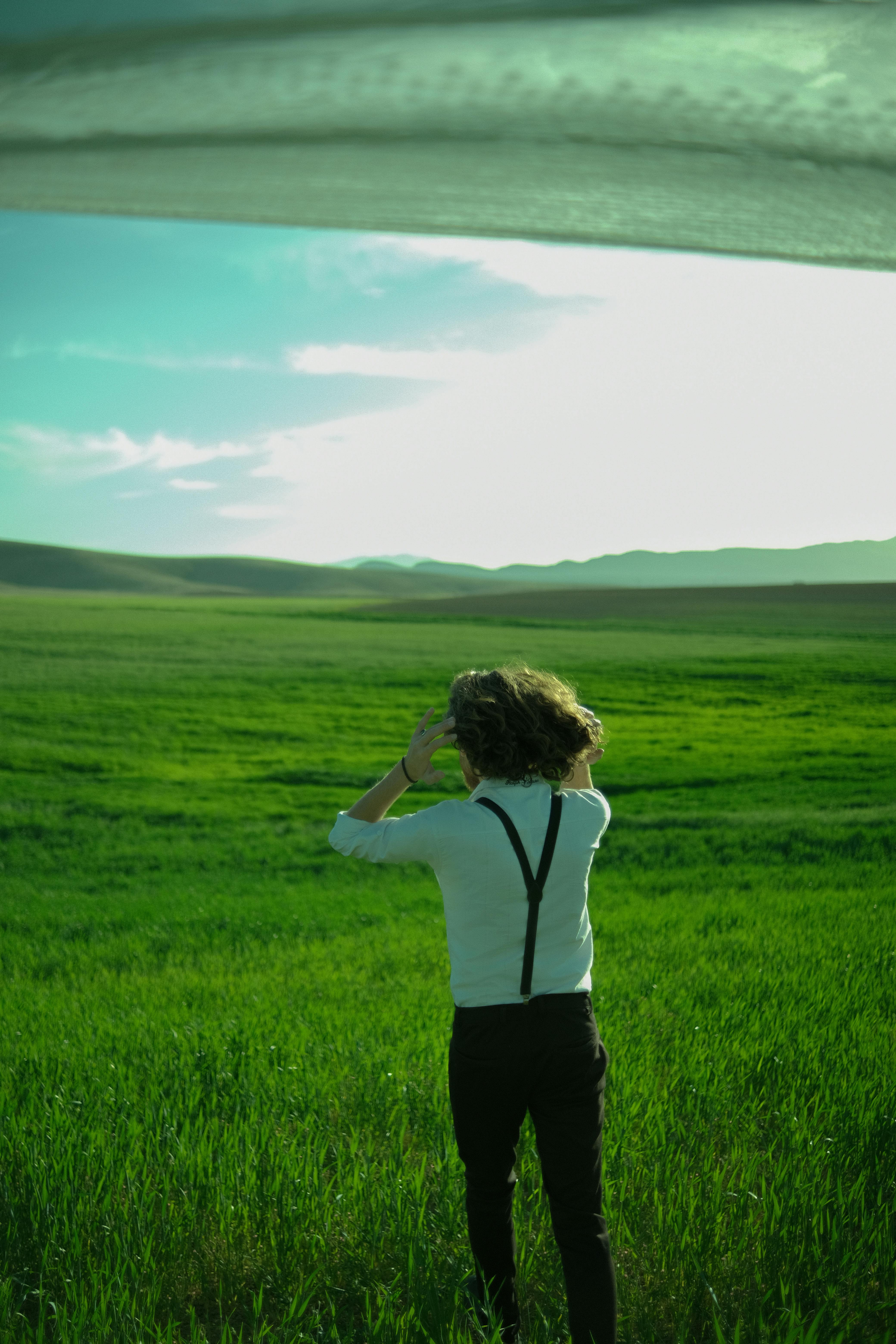 A man with curly hair walks through a vibrant green countryside landscape under a bright summer sky.