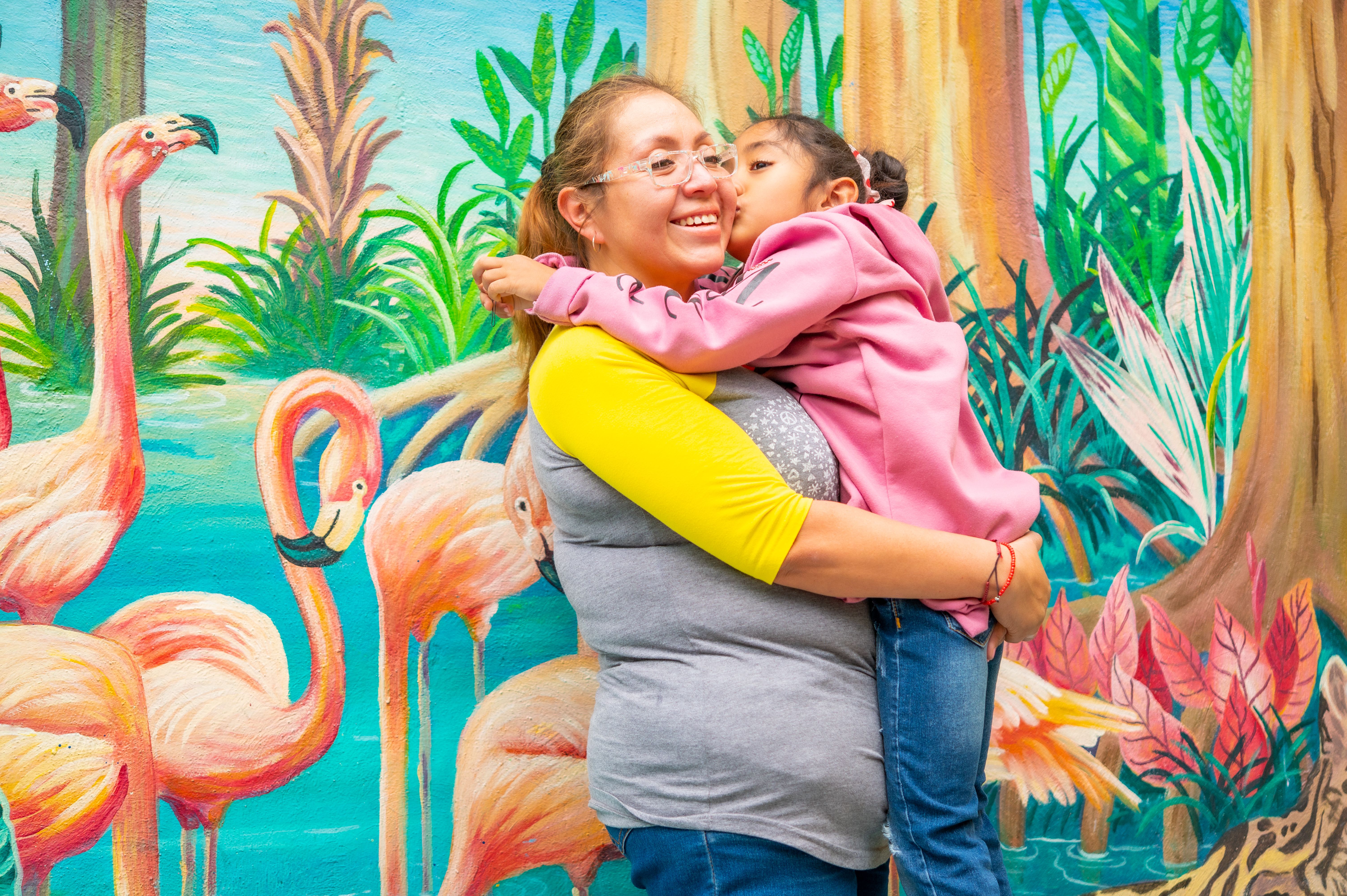 A joyful mother and daughter embrace in front of a vibrant flamingo mural