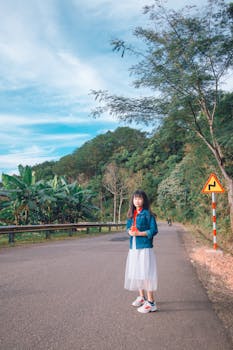 一位亚洲女子在绿树成荫的蜿蜒道路上优雅地摆着姿势，背景是蓝天
