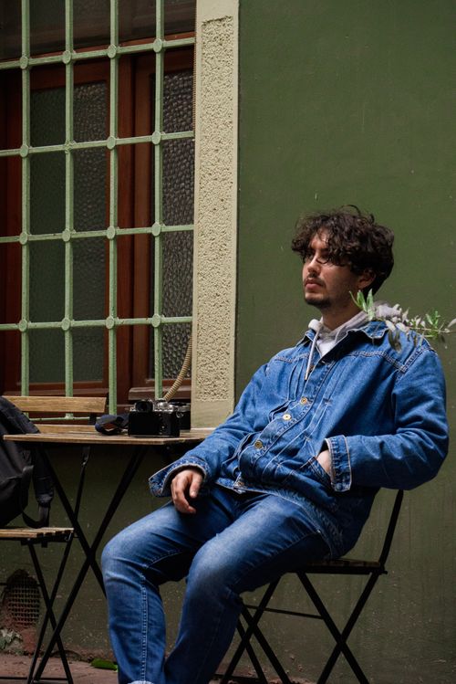 Free Man in a denim jacket and jeans sits casually on a chair outdoors, exuding a relaxed vibe. Stock Photo