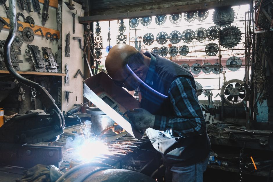 An experienced welder focused on welding in a cluttered workshop filled with tools and metal parts.