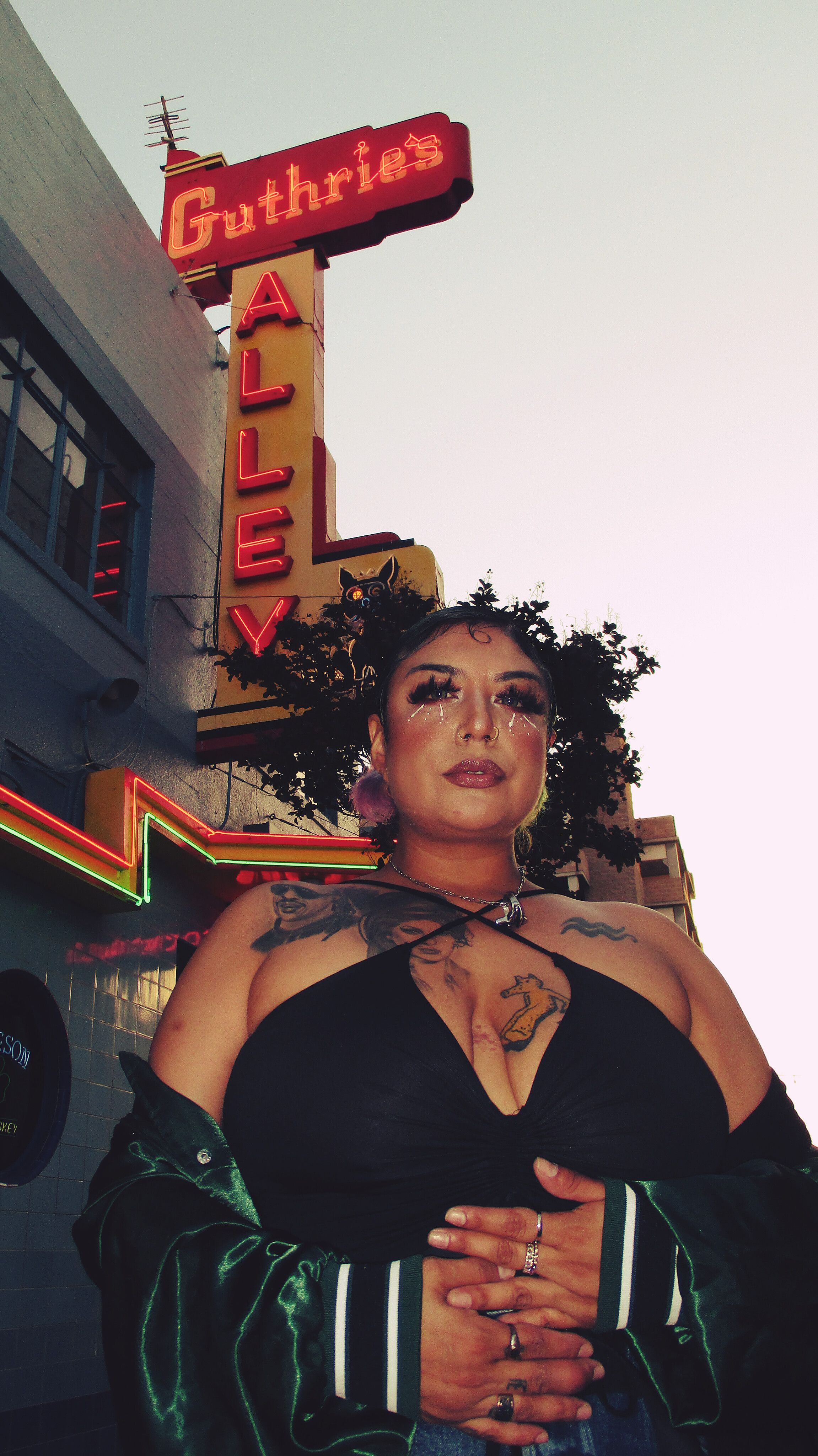 Confident woman standing under Guthrie's Alley neon sign at twilight, showcasing tattoos and fashion.