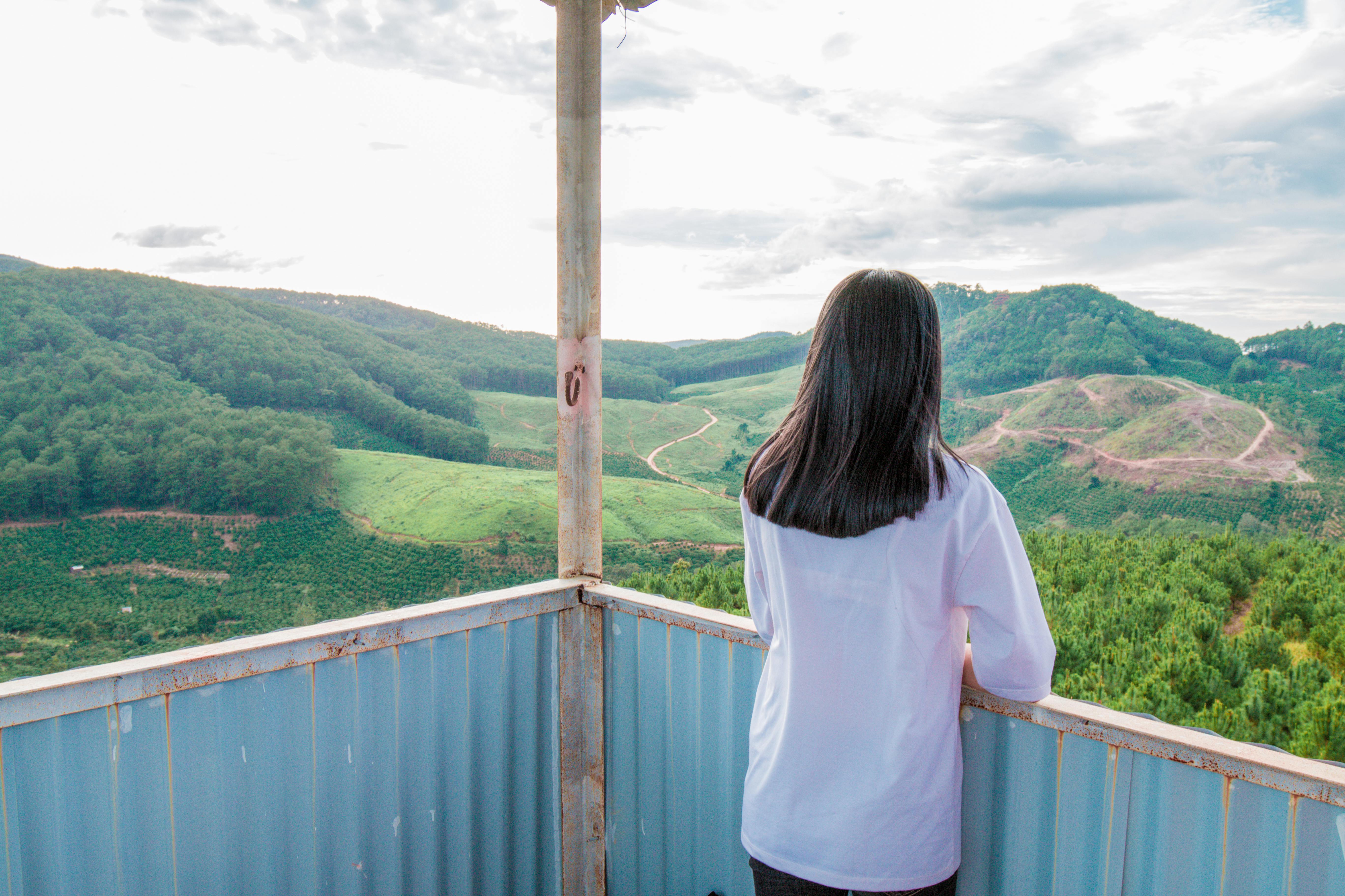 一名女子站在阳台上，凝望着明亮天空下郁郁葱葱的山地景色
