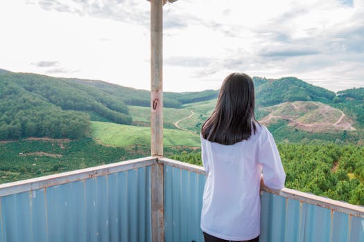 一名女子站在阳台上，凝望着明亮天空下郁郁葱葱的山地景色