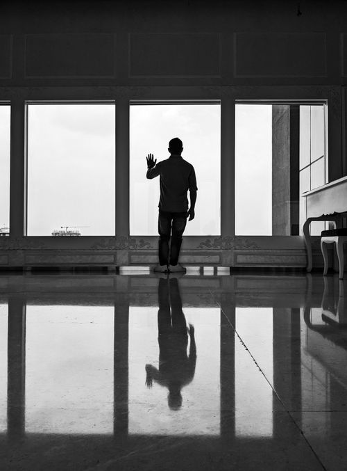 Free Silhouette of a man standing indoors, casting reflections on a shiny floor before a large window. Black and white image. Stock Photo