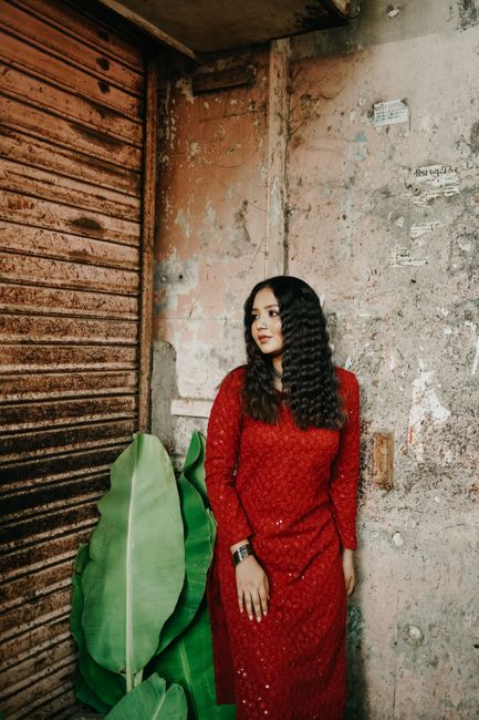 A woman in a red dress poses confidently against a textured wall with banana leaves nearby.