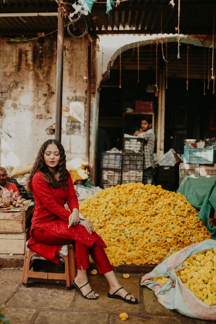 Elegant woman in a red outfit sits by a pile of yellow flowers in a bustling market.
