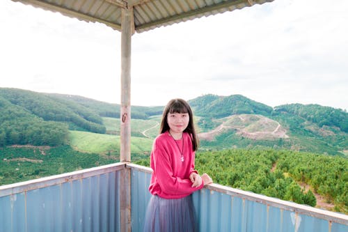 Girl Standing Beside Fence Near Mountain Under White Clouds and Blue Skies