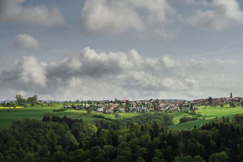 Free Beautiful countryside village surrounded by vibrant green fields and forests under a partly cloudy sky. Stock Photo