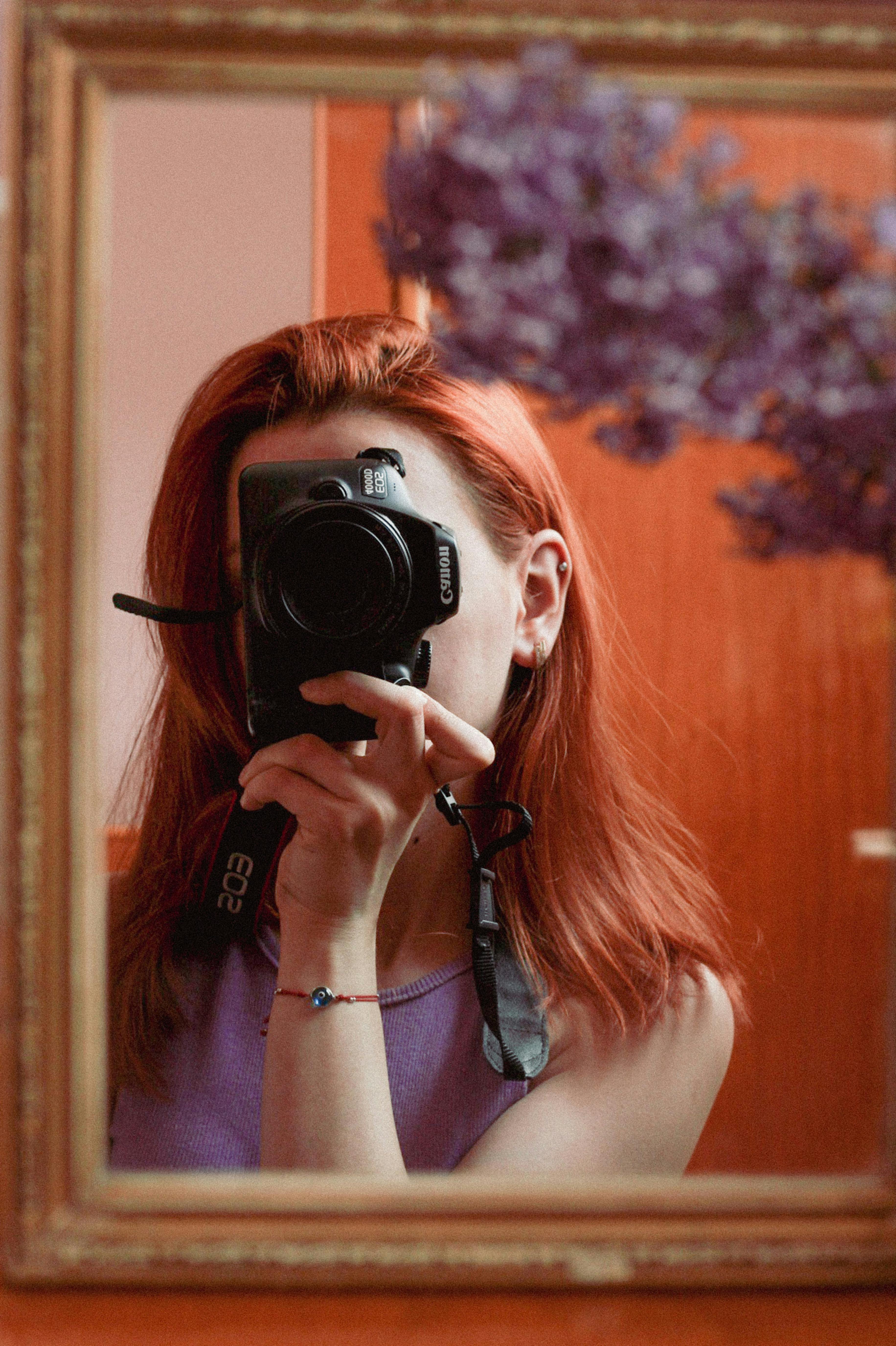 Red-haired woman takes a self-portrait with a camera in a mirror reflection.