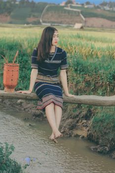 一位身着传统服装的年轻女子坐在小溪边，眺望着远处的稻田和山脉