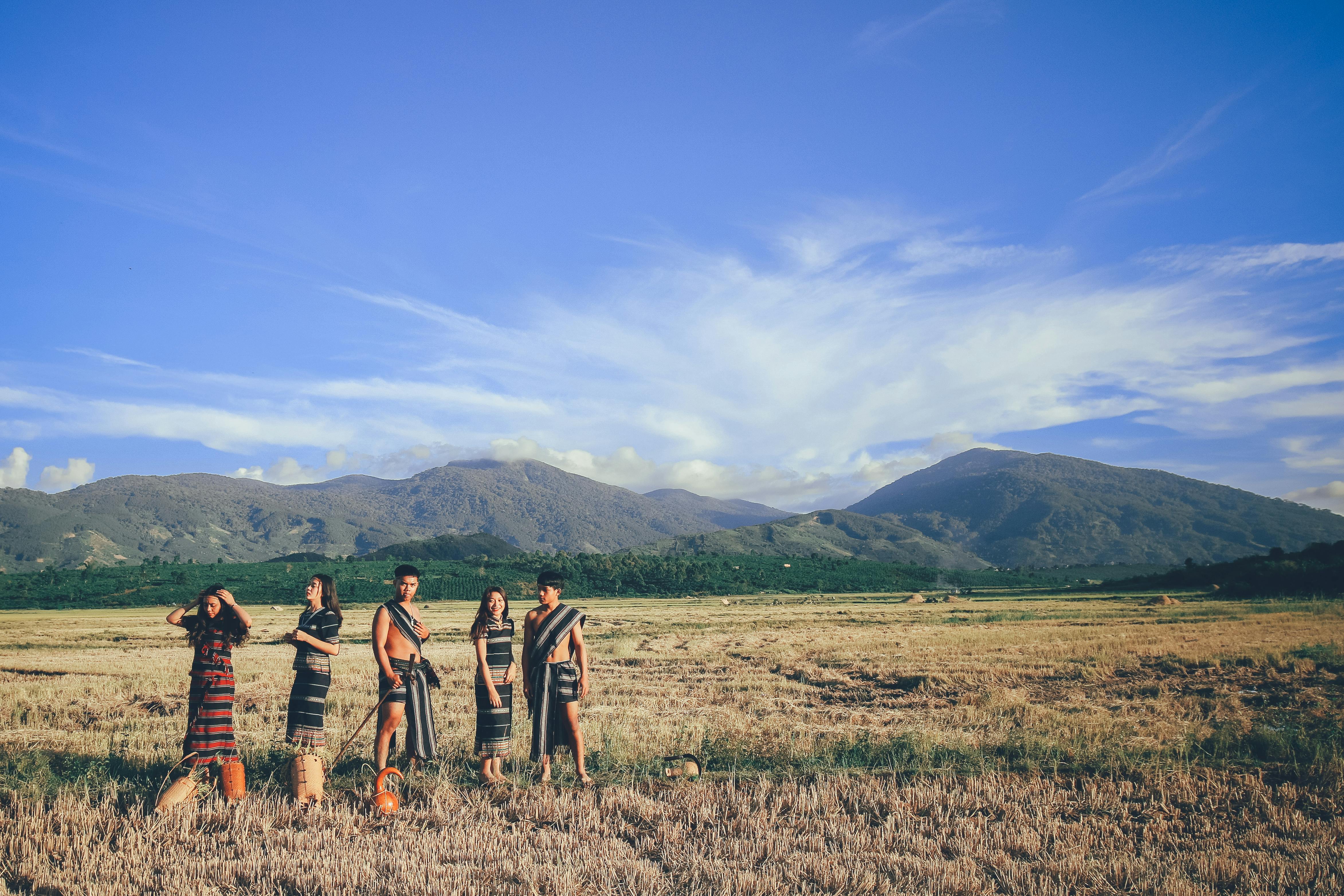 five people wearing native apparel standing on brown grassfield under blue and white cloudy sky