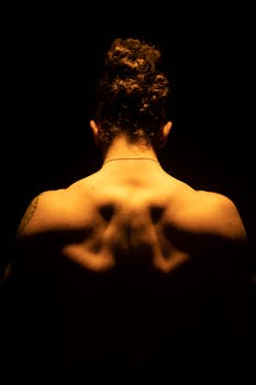 A shirtless man with muscular shoulders posing under dramatic lighting, showcasing defined back muscles.