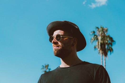 一名戴着太阳镜和帽子的时尚男子，站在明亮的蓝天下，身旁有棕榈树