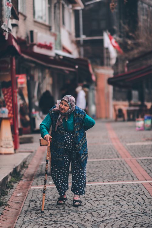 Free An elderly woman with a walking stick strolls down a charming cobblestone street. Stock Photo