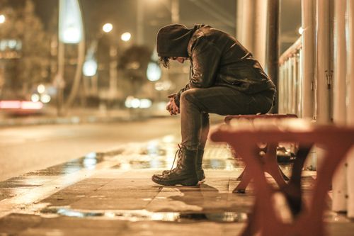 Free Man in hoodie sitting on bench at night, reflecting in rainy urban setting. Stock Photo
