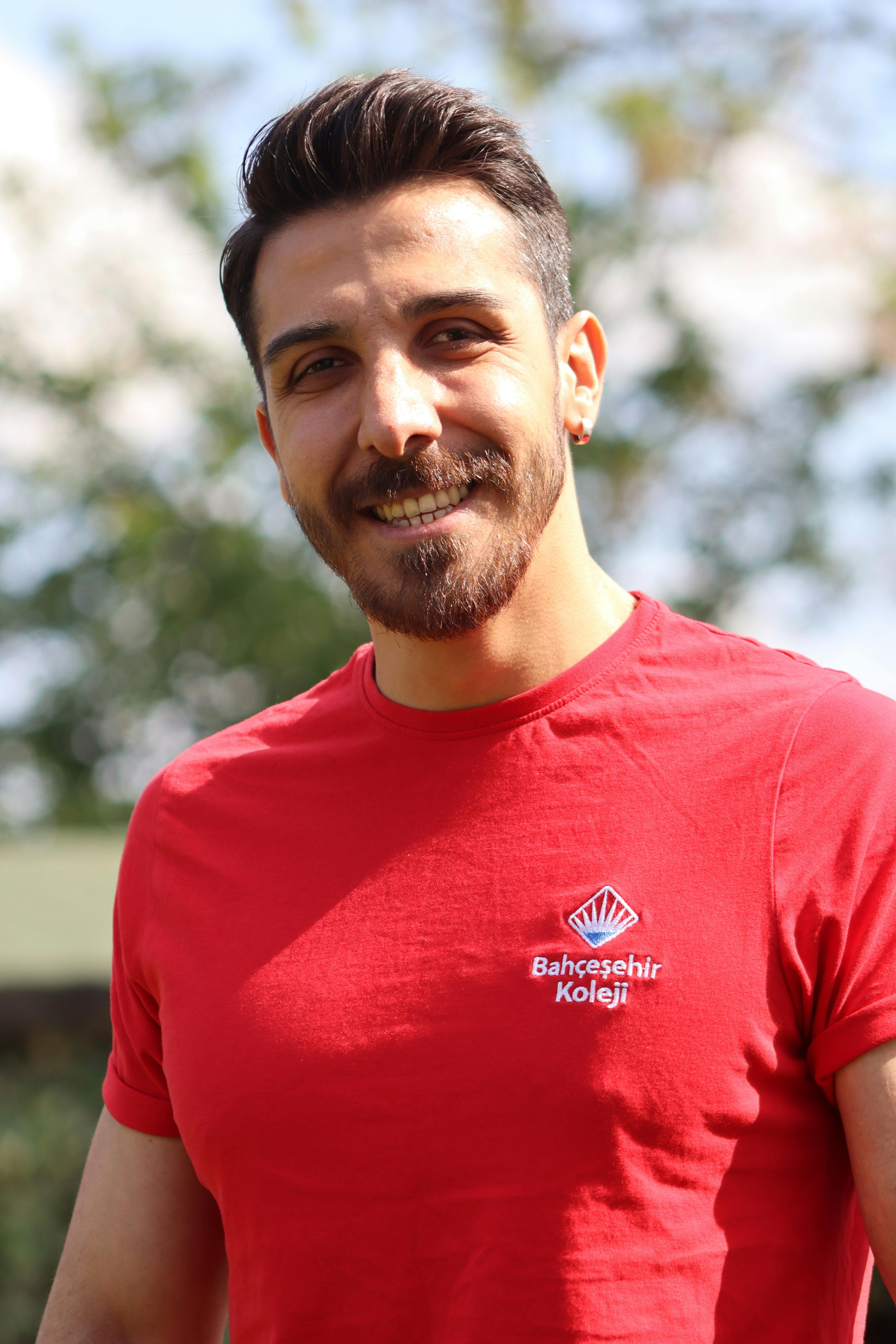 Close-up portrait of a smiling man wearing a red Bahcesehir Koleji T-shirt, standing outdoors.