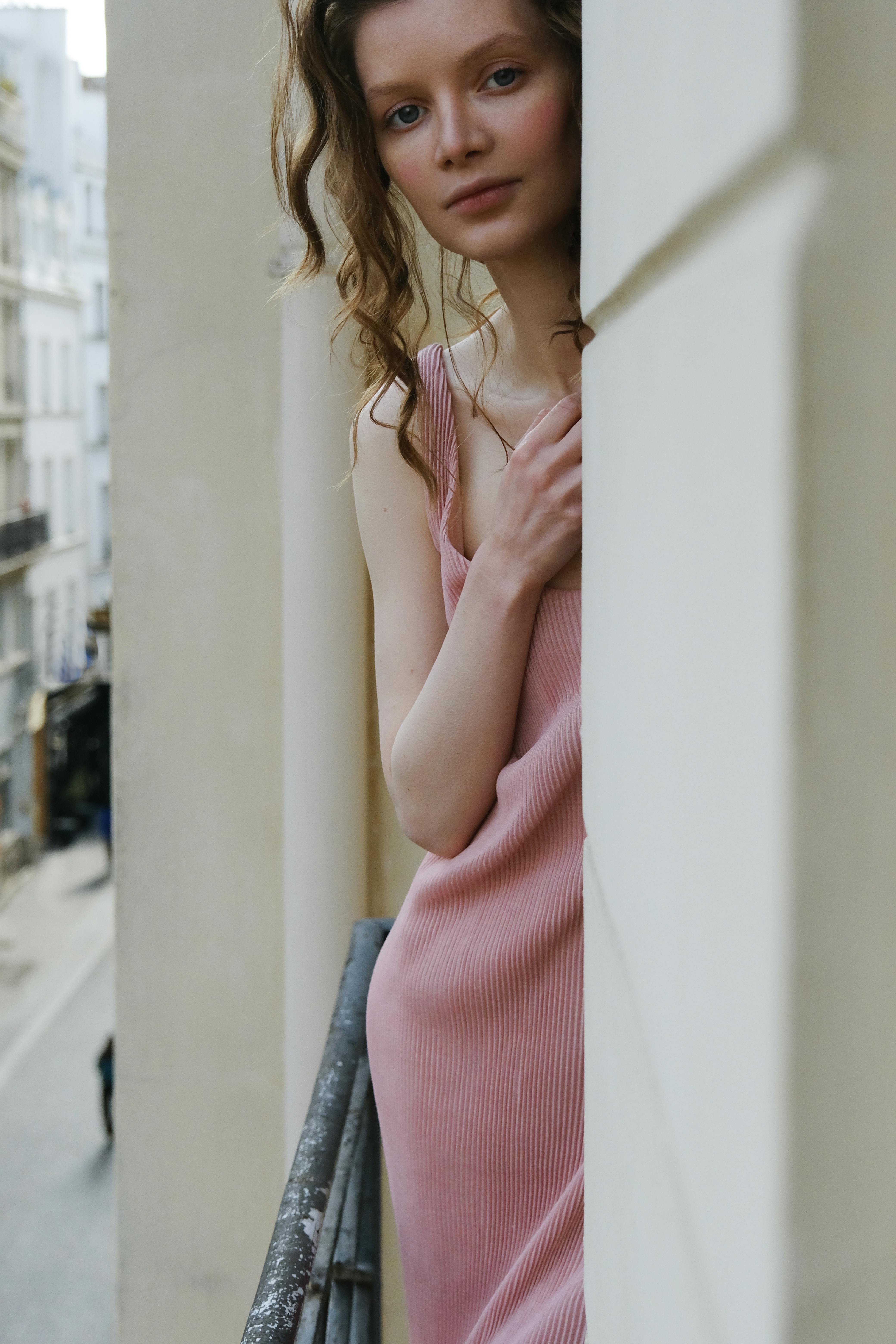Young woman in a stylish pink dress standing elegantly on a city balcony.